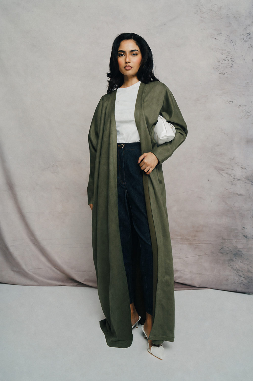 Woman wearing a long olive green coat over a white top and black pants against a gray background